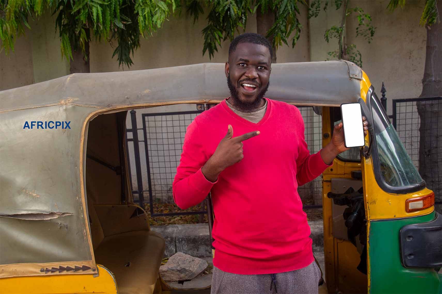 A happy African tricycle rider, also known as Keke or Maruwa, Keke rider, standing with his tricycle behind him, His is pointing at his phone.